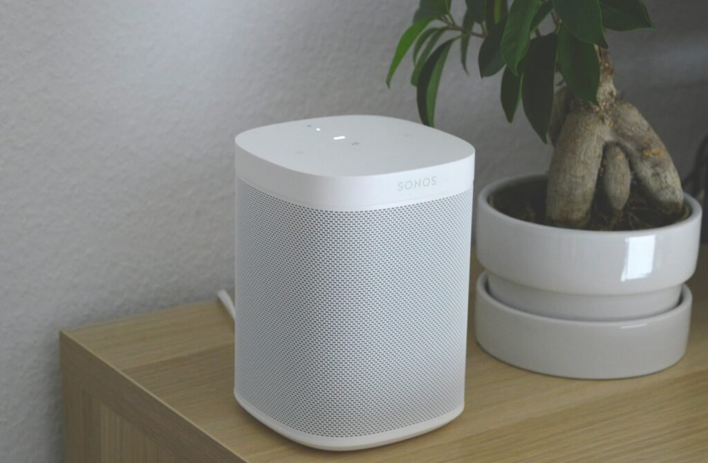 Image of a sleek, cylindrical smart speaker with a glowing LED ring at the top, indicating it is active. The speaker is compatible with both Alexa and Google Assistant, offering voice-controlled functionality for various smart home tasks.